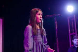 Photo of Annika Franklin, an 11-year-old girl standing in front of a microphone on a floor stand. She is singing and wearing a knee-length purple pleated dress with long ruffled sleeves.